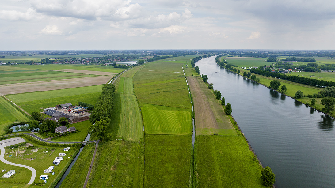 Dronefoto van de Genderensche uiterwaard die uit een groot stuk grasland bestaat. De uiterwaard grenst links aan een boerderij en rechts aan de Bergsche Maas (dezelfde foto als boven maar vanaf de andere kant genomen).
